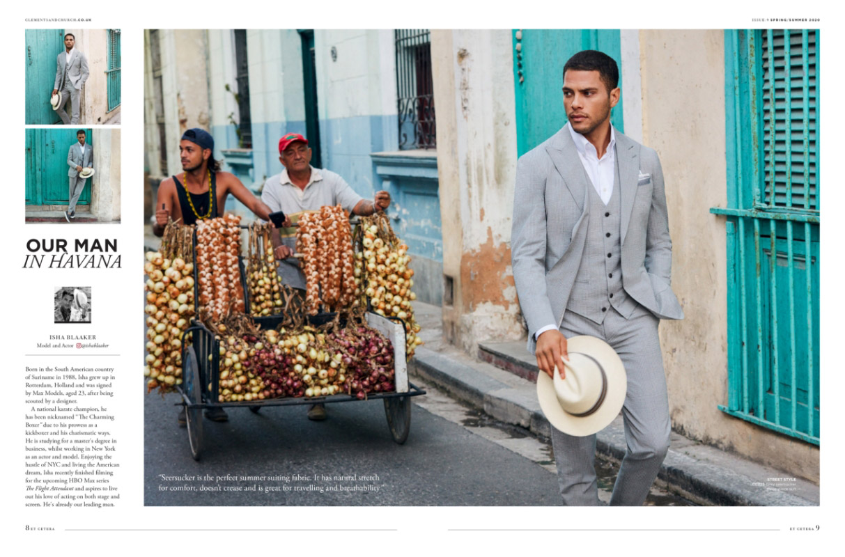 Man wearing a grey searsucker suit and holding a Panama hat walking down a street in Havana